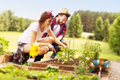 The image depicts a woman and a man working together in a well-maintained garden with raised bed planting areas. The woman, wearing a white t-shirt, patterned shorts, and a red headband, is actively tending to young plants with a small gardening tool, while the man, dressed in a plaid shirt and straw hat, observes and assists. The garden features rich, dark soil and healthy green foliage, with several small plants and seedlings growing in the beds. In the foreground, a decorative garden figure resembling a yellow bird is visible, alongside a small garden ornament with colorful sections. The background reveals a lush, expansive landscape with a neatly trimmed lawn, dense trees, and a clear sky, suggesting a peaceful outdoor environment on a sunny day. This scene exemplifies the benefits of professional gardening services such as planting, garden maintenance, and outdoor landscaping, consistent with the services offered by Lawn Mowing Cranford in the local area.