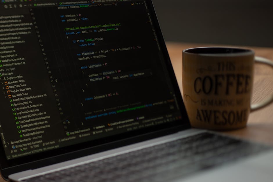 A close-up view of a laptop displaying lines of code on its screen, with a ceramic coffee mug positioned slightly behind it. The mug has a beige background with black and dark red text that reads 'Thi